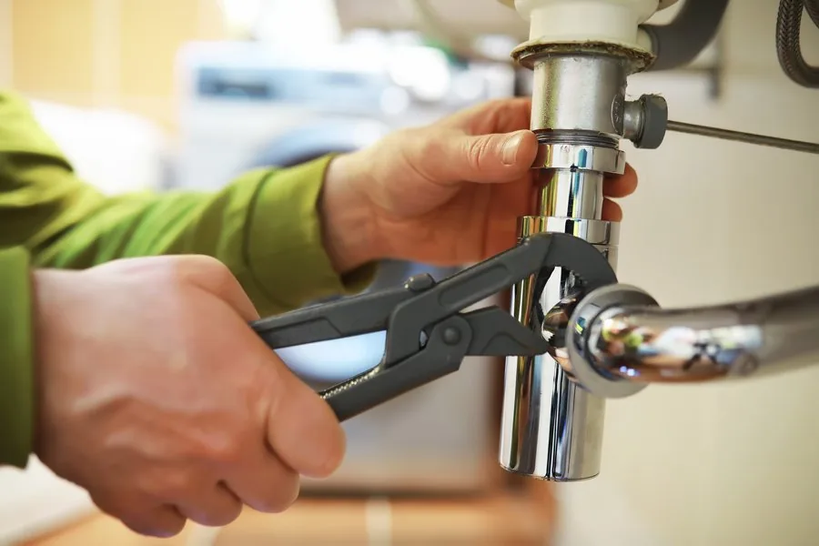 A man in overalls stands beside a water heater, holding a wrench, ready for maintenance or repair work.