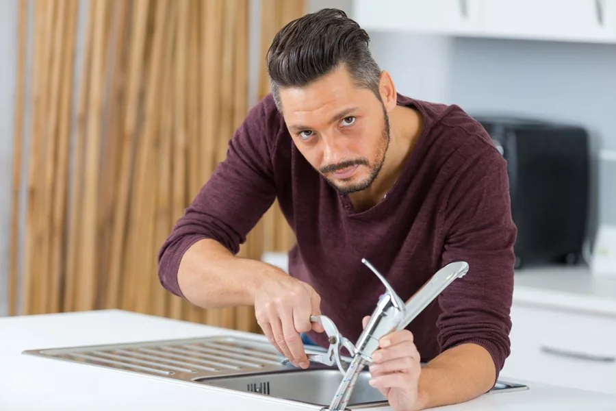 A man is diligently repairing a kitchen sink, focused on his task with tools and plumbing parts nearby.

