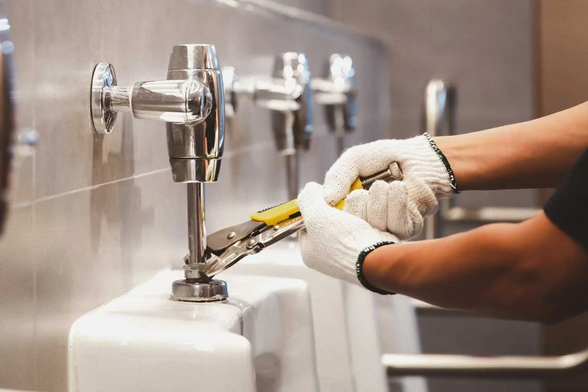 A person wearing white gloves is repairing a urinal in a restroom setting.

